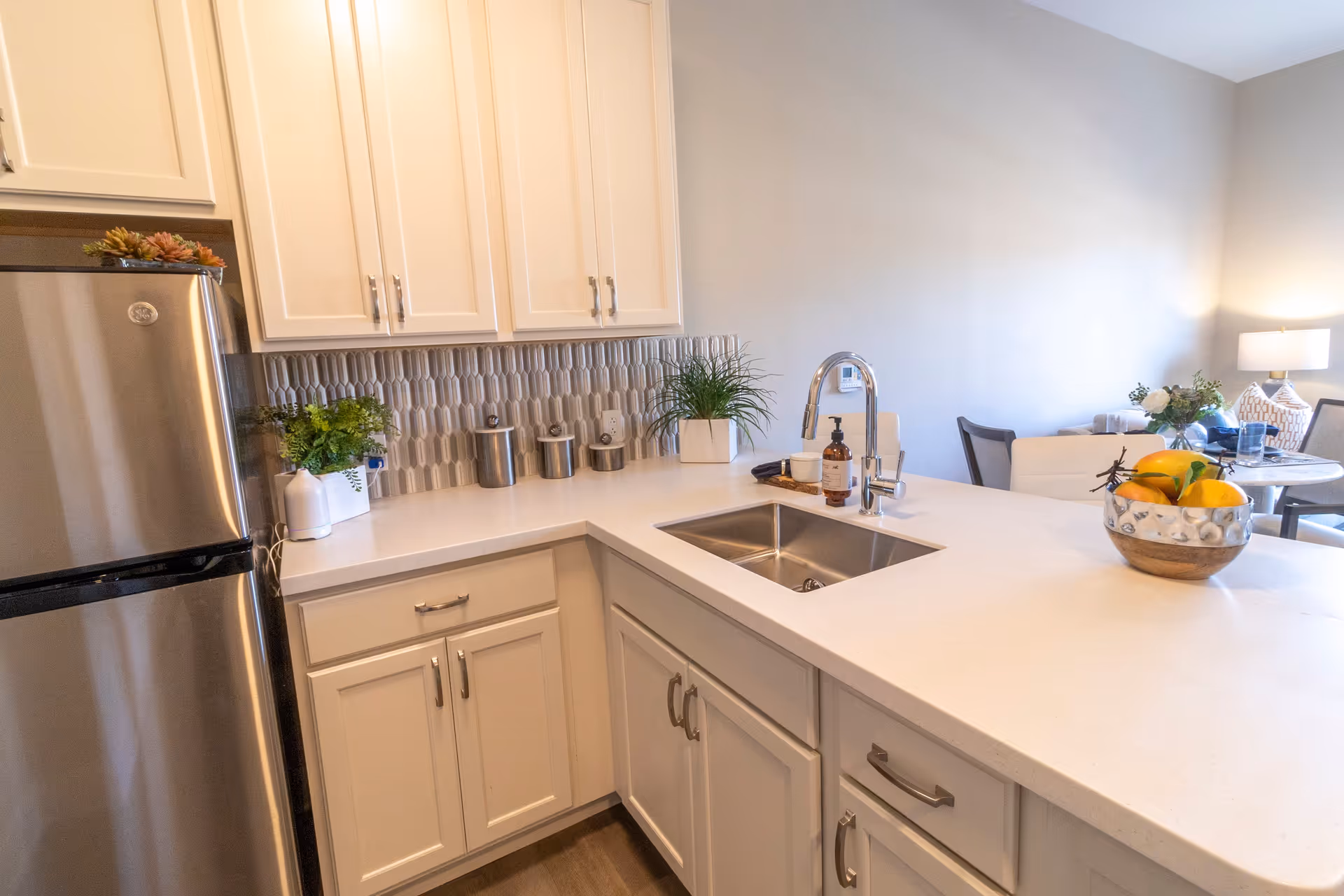 Modern kitchen area with white cabinets, a stainless steel refrigerator, a white countertop with a built-in sink and chrome faucet, decorative plants, a soap dispenser, and a bowl of fruit. In the background, a dining table with chairs and a lamp are visible.