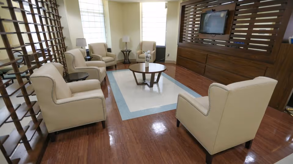 A cozy living room area with six beige armchairs arranged around a round wooden coffee table on a floor with a white and blue patterned section. The room features wooden flooring, a wooden slatted partition, two windows with blinds, two side tables with lamps, and a wall-mounted flat screen TV on a wooden panel.