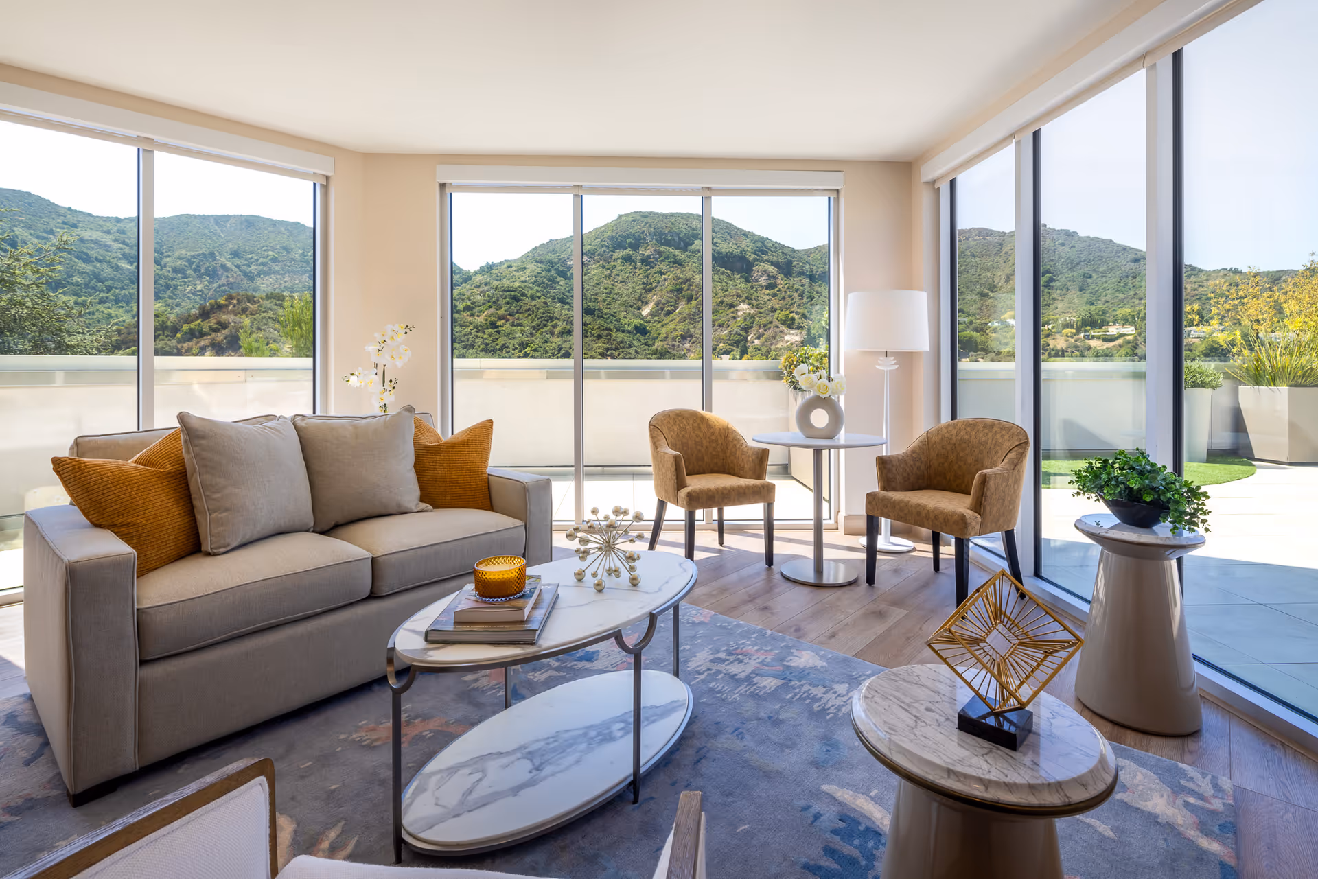 Bright living room with large floor-to-ceiling windows showing a scenic mountain view. The room features a beige sofa with orange and gray cushions, two brown armchairs around a small round table with a vase of flowers, a floor lamp, a marble coffee table with decorative items, and a patterned rug on wooden flooring.