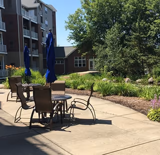 Sunny courtyard with round patio tables and chairs under blue umbrellas, landscaped beds, and a multi-story building in the background.