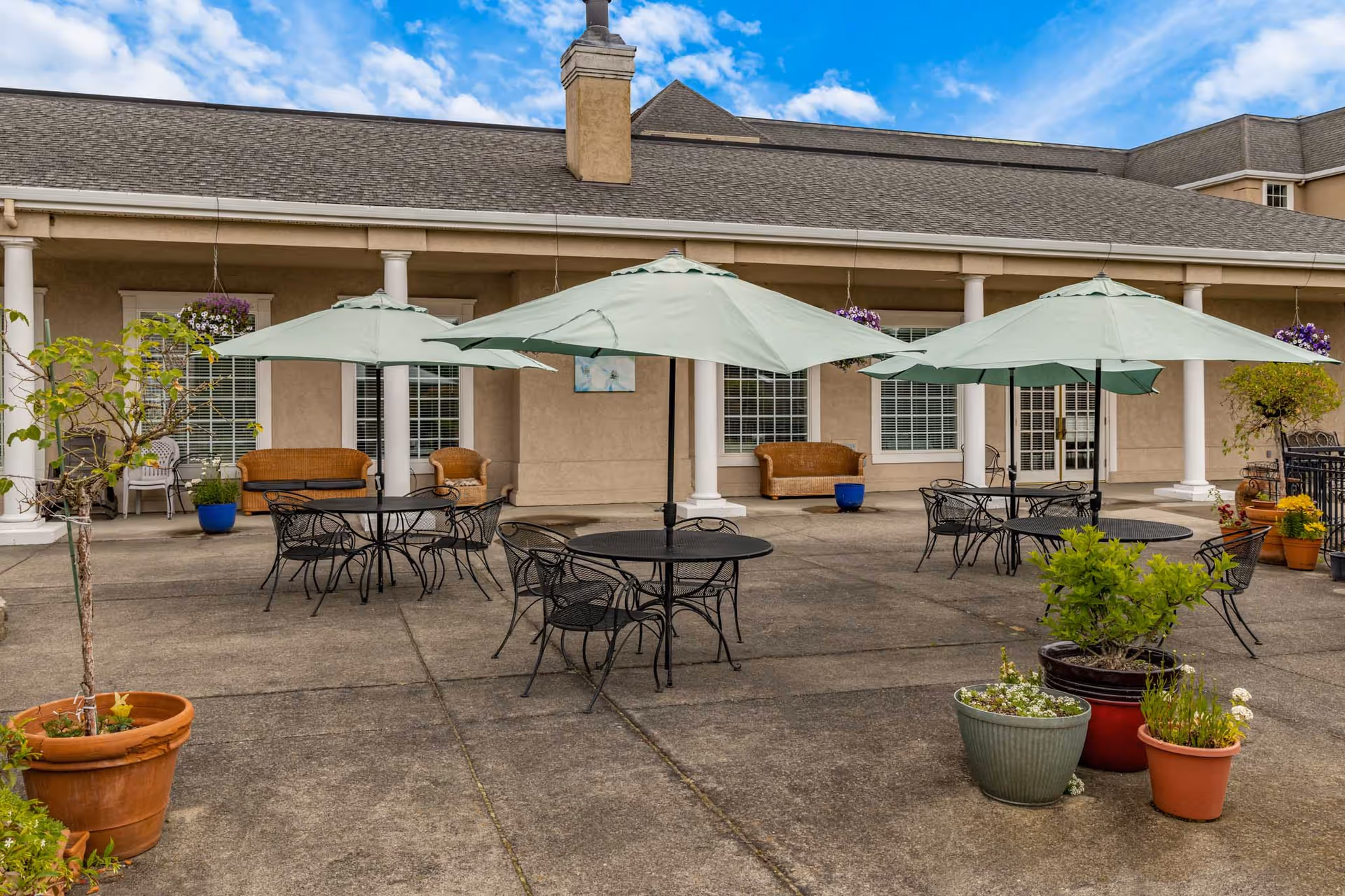 Outdoor patio with round metal tables, green umbrellas, chairs, and potted plants in front of a building.