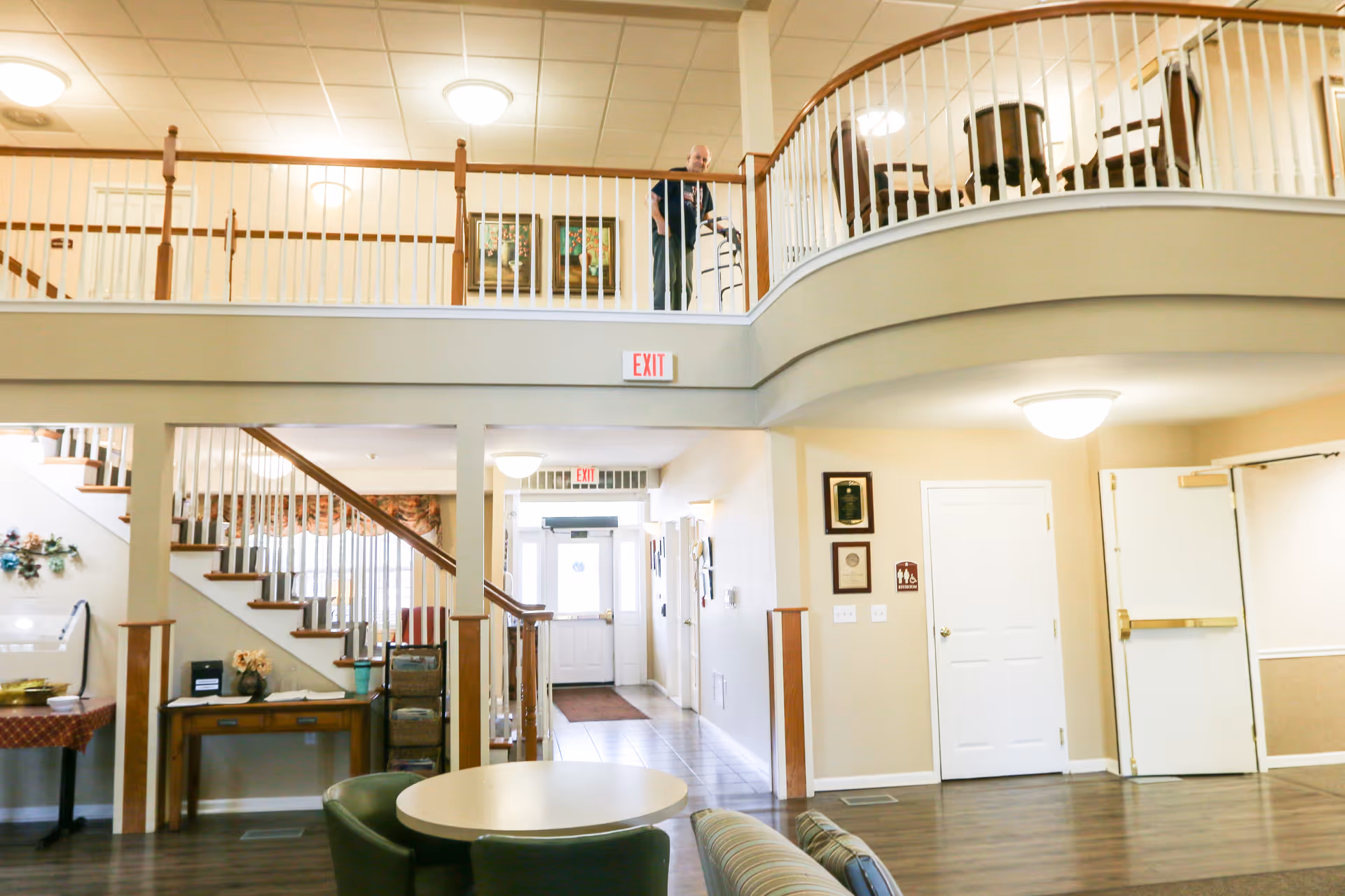Interior view of a senior living facility lobby area with a staircase leading to an upper floor balcony. A person with a walker is standing on the balcony. The space includes a round table with chairs, a couch, and various wall decorations. There are exit signs above doors and soft lighting from ceiling fixtures.