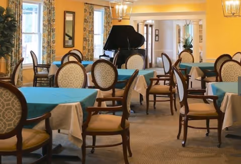 A dining room with several tables covered with blue tablecloths and beige chairs with patterned backs. The room has yellow walls, large windows with floral curtains, and a black grand piano in the background.