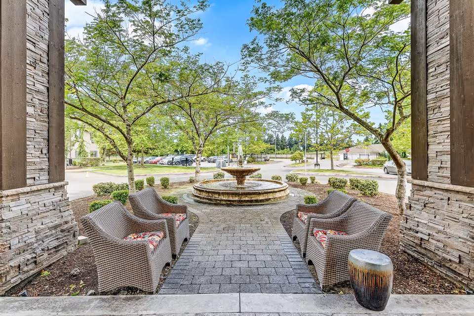 Outdoor seating area with four wicker chairs featuring floral cushions arranged around a paved pathway leading to a circular stone fountain. The area is framed by two stone pillars and surrounded by trees and shrubs under a blue sky with scattered clouds.