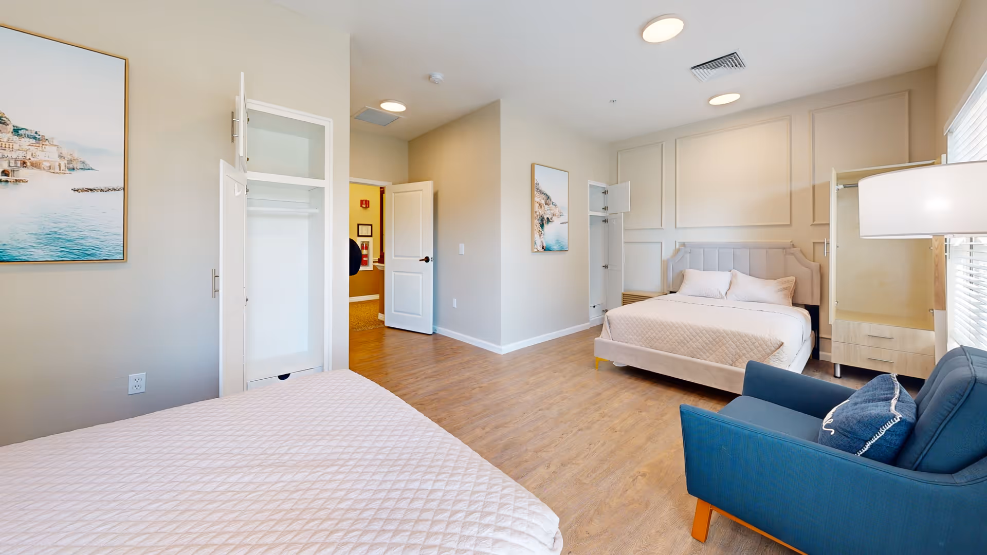 A bright bedroom with two beds covered in light-colored quilts, a blue armchair with a decorative pillow, two open closets, a large window with blinds, and framed coastal artwork on the walls. The room has wood flooring and soft beige walls with decorative molding.