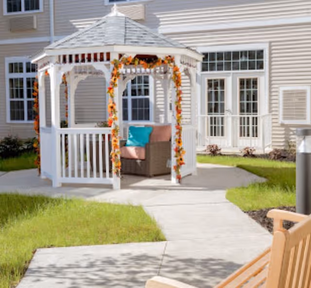 A white gazebo decorated with garlands sits on a paved courtyard with cushioned seating in front of a beige assisted living building.