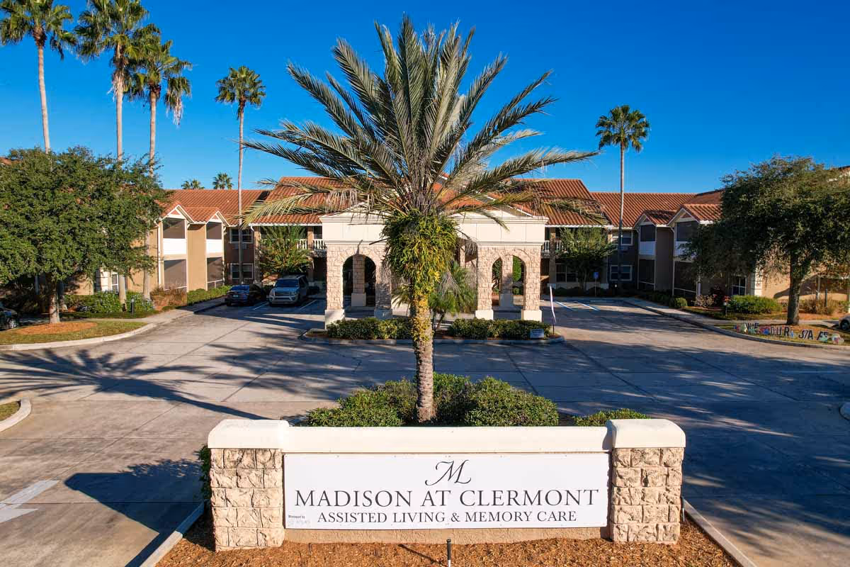 Exterior view of Madison At Clermont assisted living and memory care facility with a stone entrance sign, palm trees, and a two-story building with a red-tiled roof under a clear blue sky.