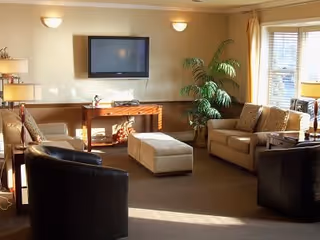 A cozy living room with beige walls and carpet, featuring a wall-mounted flat-screen TV above a wooden console table. The room has two beige sofas with patterned cushions, two black leather armchairs, a beige ottoman in the center, a tall green plant near the window, and soft lighting from wall sconces and a floor lamp.