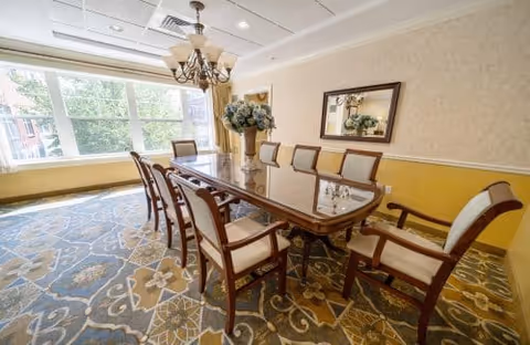 A bright dining room with a large wooden dining table surrounded by eight upholstered chairs. A chandelier hangs above the table, and a large window lets in natural light. The room has patterned carpet, yellow and cream walls, and a mirror with a floral arrangement beneath it on one wall.