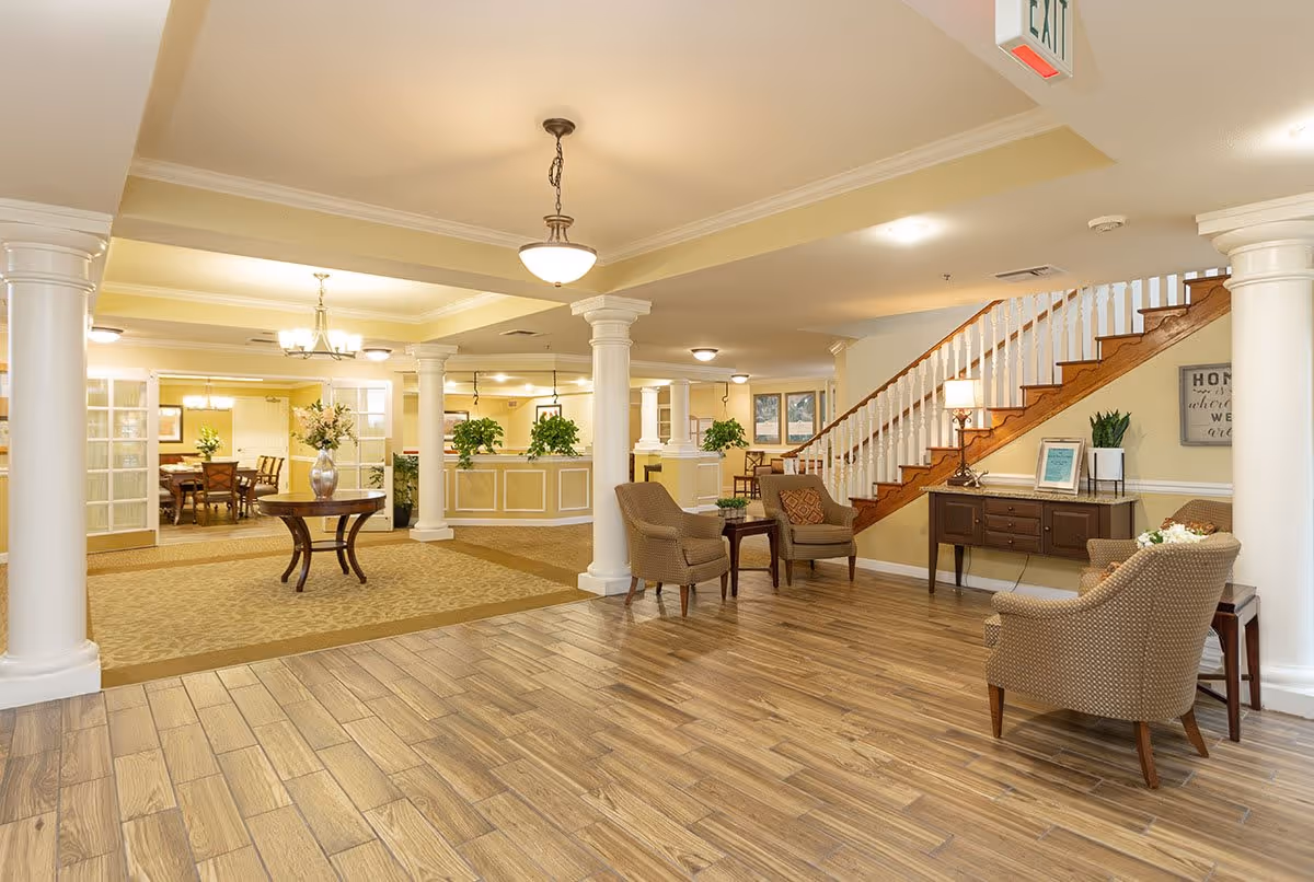 A spacious and well-lit senior living facility common area with wooden flooring, beige walls, and white columns. The room features comfortable armchairs arranged around small tables, a wooden staircase with white railings, a sideboard with a lamp and framed pictures, and a round table with a vase of flowers. In the background, there is a dining area with a chandelier and a table with chairs.