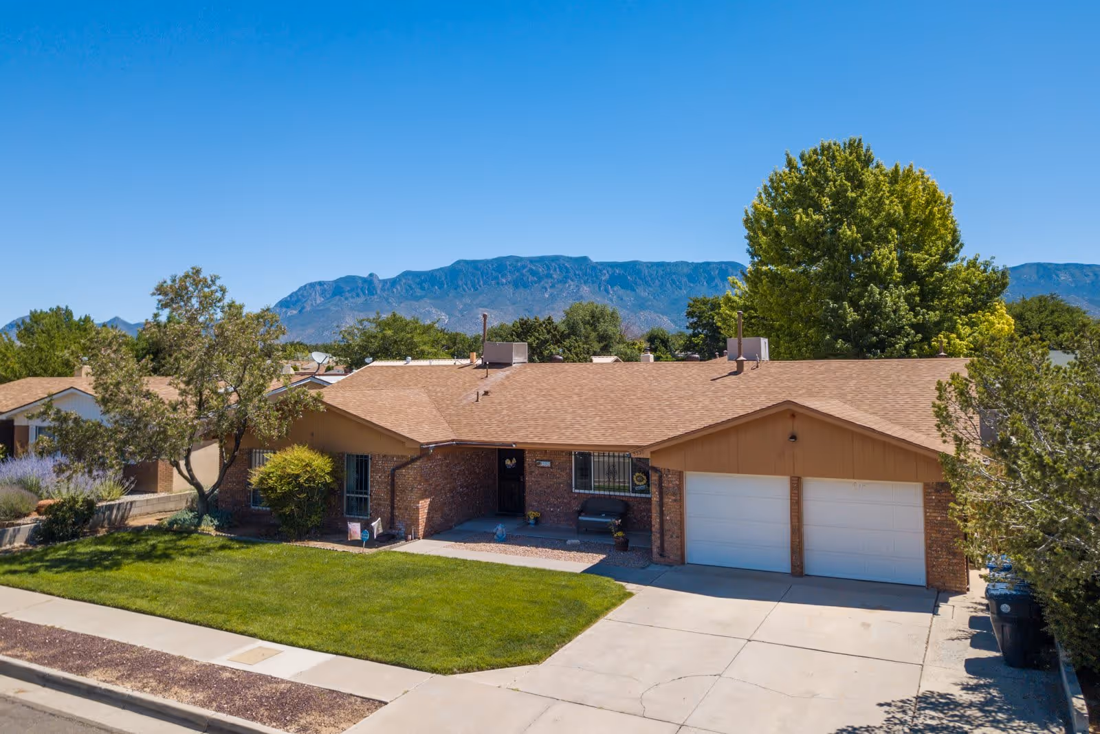 Single-story brick residential building with a two-car garage, green lawn, trees, and a mountain range in the background under a clear blue sky.