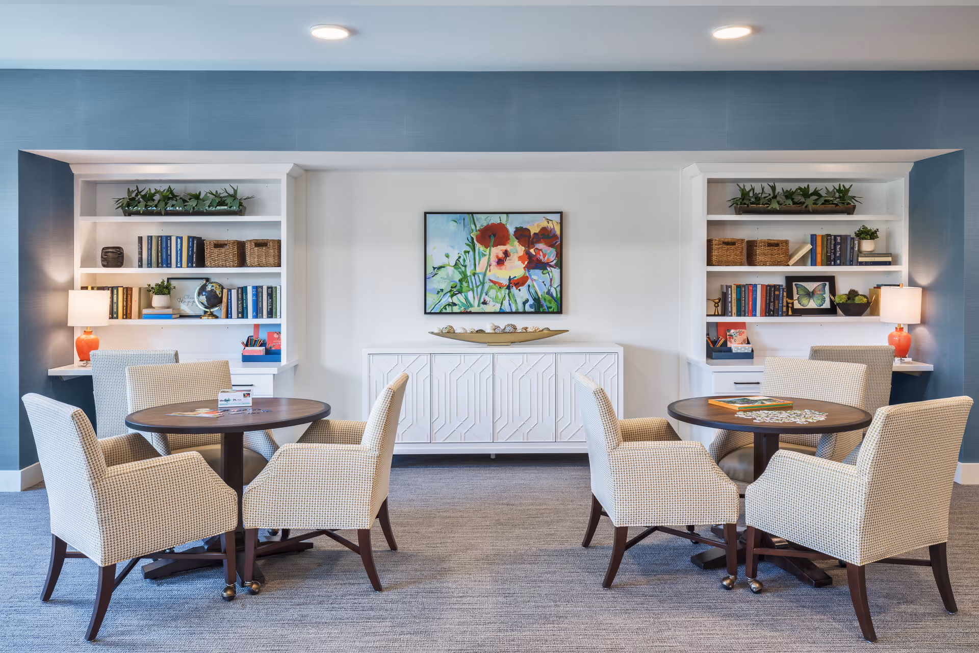 A cozy common area with two round wooden tables, each surrounded by four beige upholstered chairs. Behind the tables are built-in white bookshelves filled with books, baskets, plants, and decorative items. A white cabinet with a decorative bowl on top is centered between the bookshelves, and a colorful floral painting hangs above it. The walls are painted blue with a white accent wall in the middle, and the floor is carpeted in a neutral tone.