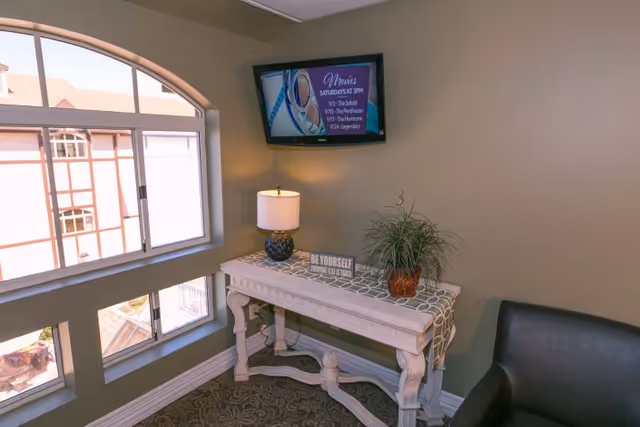 A cozy corner in an assisted living facility with a decorative wooden table holding a lamp, a small potted plant, and a sign that reads 'BE YOURSELF Everyone else is taken.' Above the table, a wall-mounted TV displays a movie schedule. There is a large window with an arched top letting in natural light, and a dark armchair is partially visible on the right.