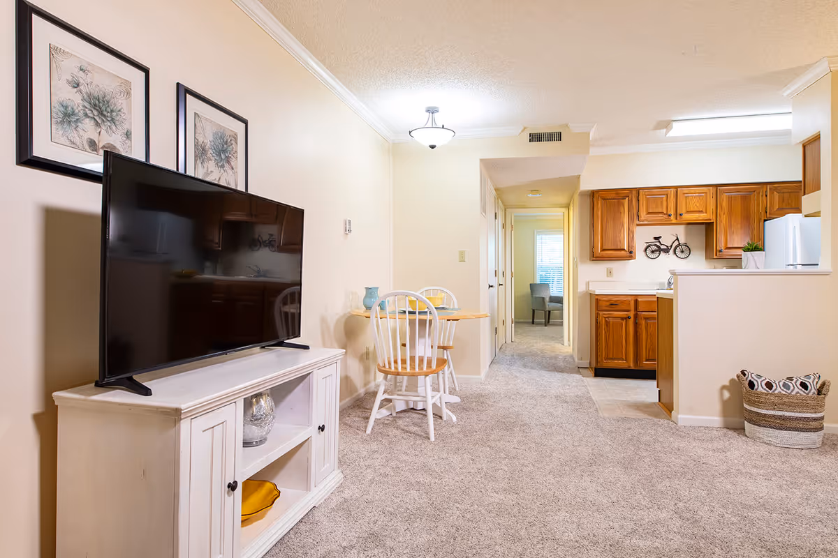 Open living area with a TV on a white cabinet, a small dining table, and a kitchen with wooden cabinets in the background.