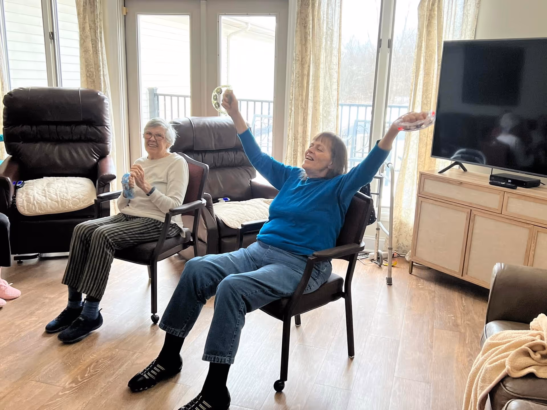 Two elderly women sitting on chairs in a bright living room with large windows and curtains. One woman is smiling and holding a water bottle, while the other is raising her arms joyfully, holding tambourines. The room has wooden flooring, a TV on a wooden cabinet, and comfortable recliner chairs.