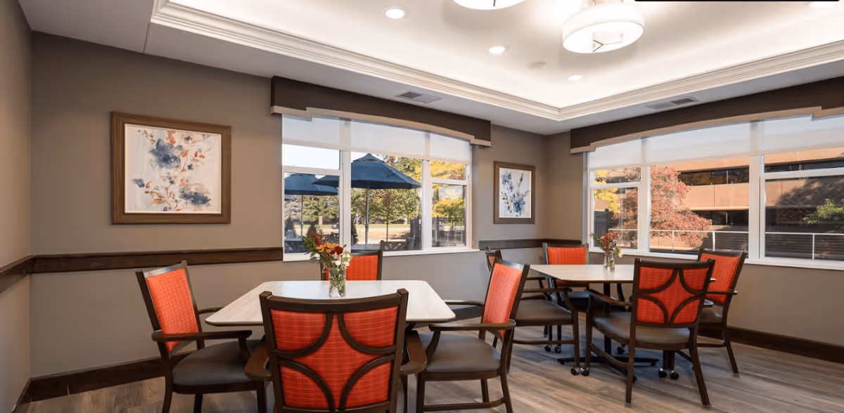 A bright dining room with two square tables, each surrounded by four chairs with red cushioned backs and brown seats. The room has large windows with white blinds, allowing natural light to enter and showing an outdoor view with trees and umbrellas. The walls are painted beige and decorated with framed floral artwork. The ceiling has recessed lighting and a modern circular light fixture.