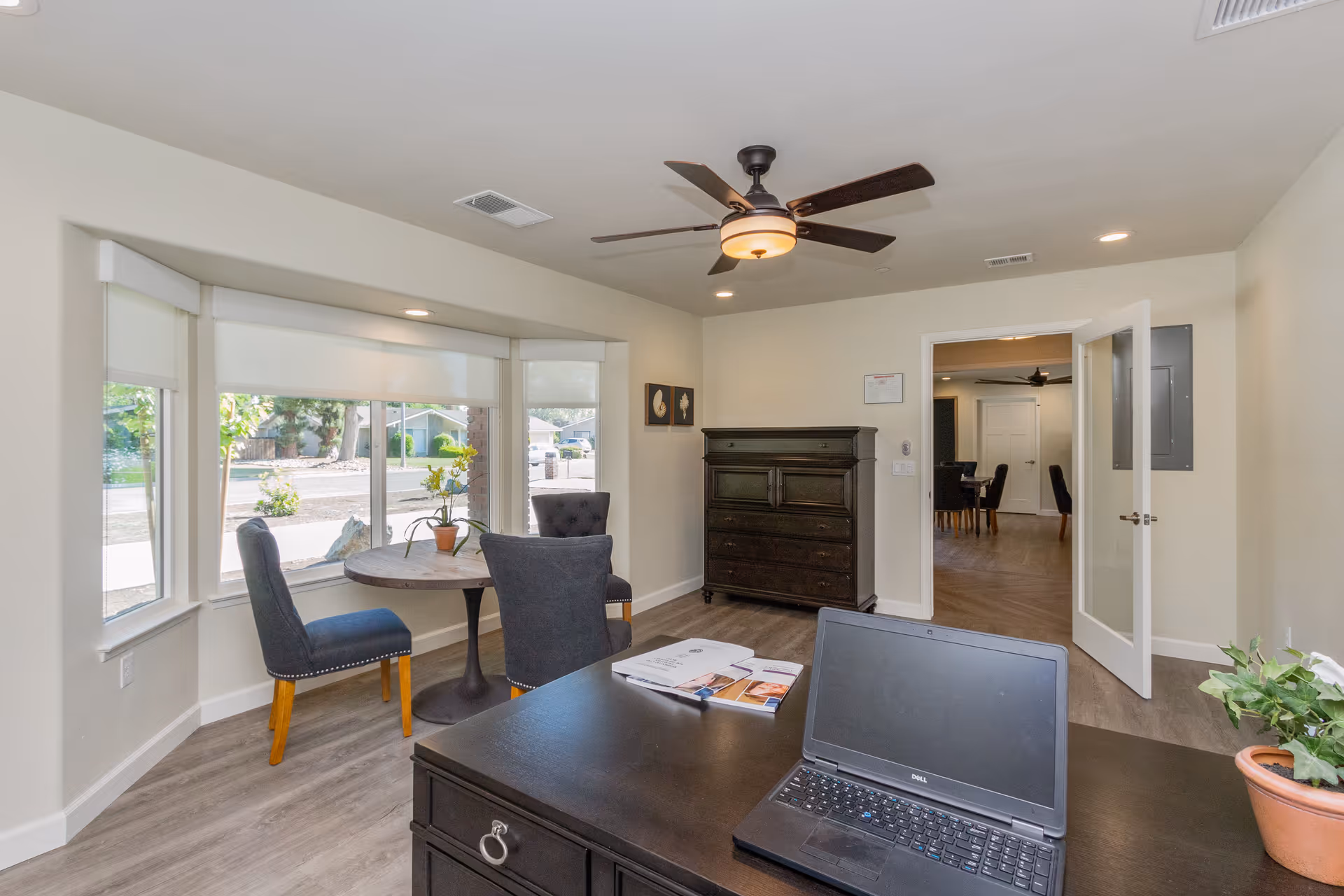 A bright office space in Calimyrna Assisted Living with a dark wooden desk featuring a laptop and open book. There is a round table with two upholstered chairs near large windows that let in natural light. A dark wooden chest of drawers is against the wall, and a ceiling fan with a light fixture is mounted on the ceiling. An open door leads to another room with additional chairs and a similar ceiling fan.