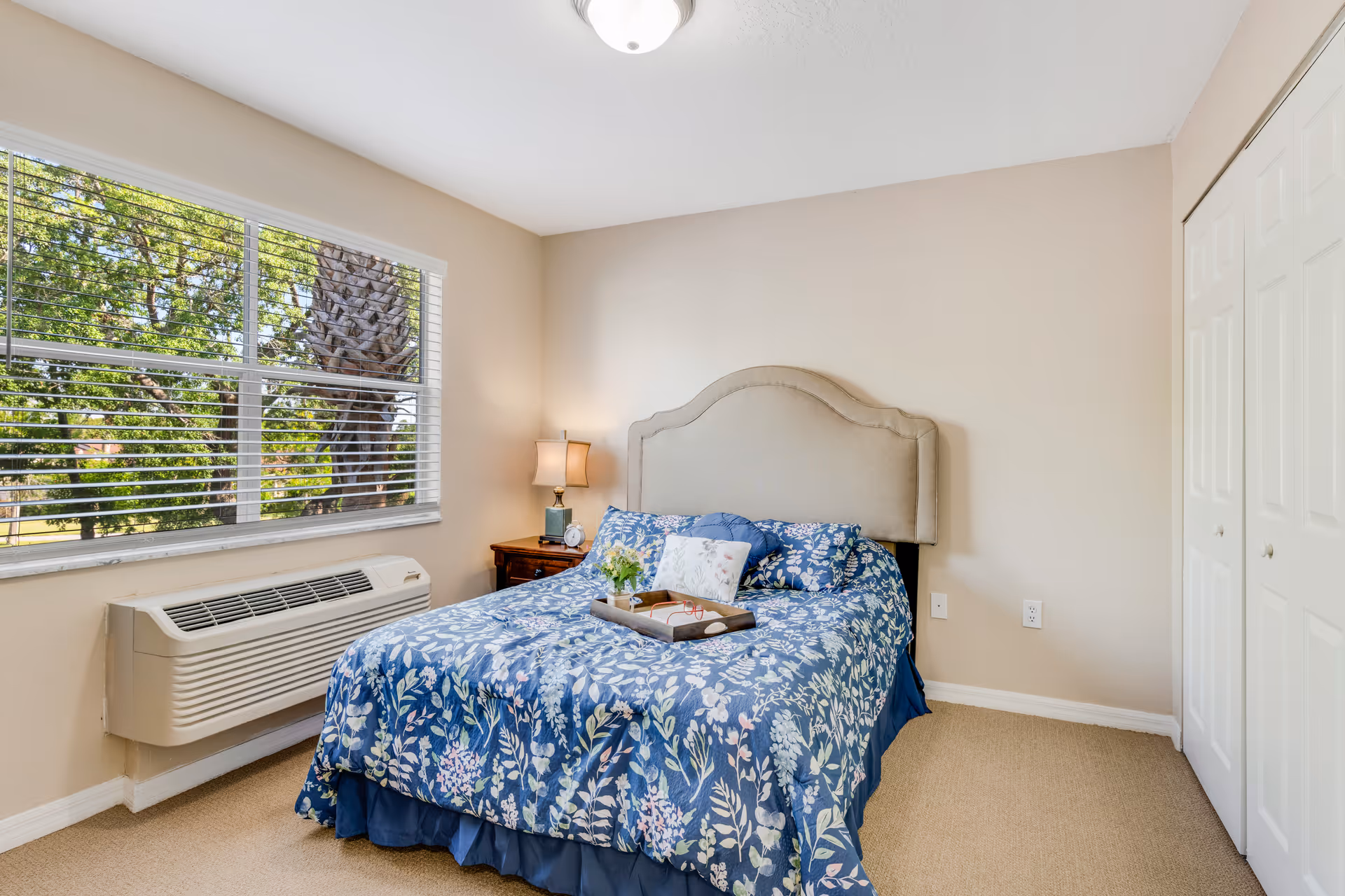 A bedroom with a bed covered in a blue floral comforter and matching pillows. There is a beige upholstered headboard, a wooden nightstand with a lamp and clock, a window with white blinds showing green trees outside, a wall-mounted air conditioning unit below the window, and a closed white closet door on the right.