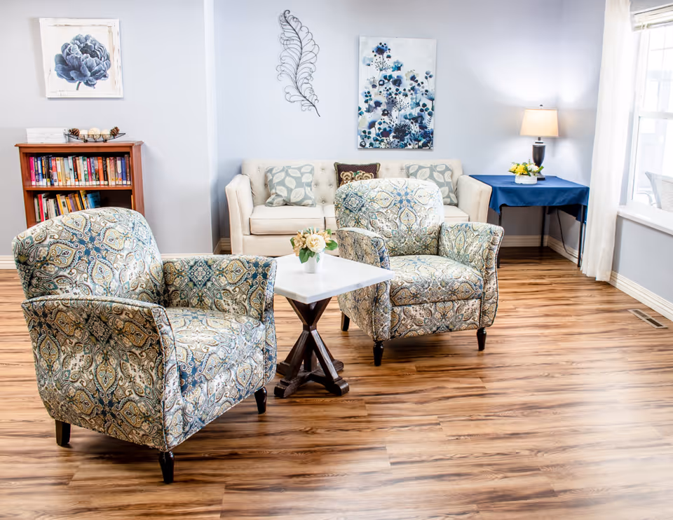 Cozy living room area with two patterned armchairs around a small table, a sofa, bookshelf, lamp, and wall art.