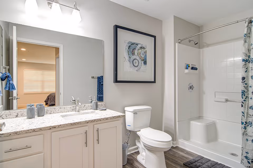 A clean and modern bathroom featuring a white vanity with a granite countertop, a large mirror above the sink, a toilet, and a shower with a white tiled wall and a floral shower curtain. A framed abstract artwork hangs on the wall above the toilet. The bathroom has light-colored walls and wood-style flooring. A glimpse of a bedroom with a window and blinds is visible through the open door.
