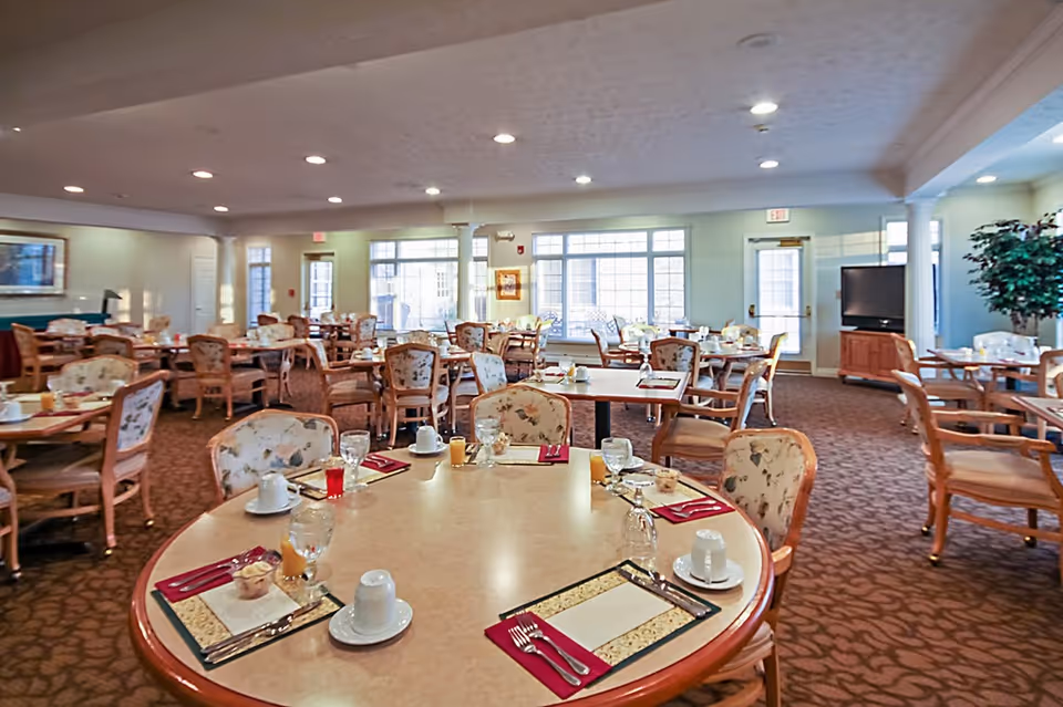A spacious dining room with multiple round and rectangular tables set with placemats, napkins, cups, glasses, and utensils. The chairs have floral upholstery, and large windows allow natural light to fill the room. There is a television on a wooden stand and a large potted plant in the corner.