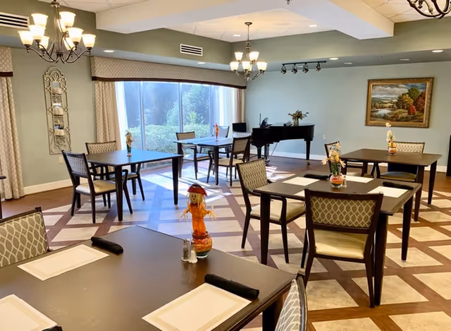 Bright dining room with multiple square tables and chairs set with placemats, chandeliers overhead, a grand piano by large windows, and wall artwork.