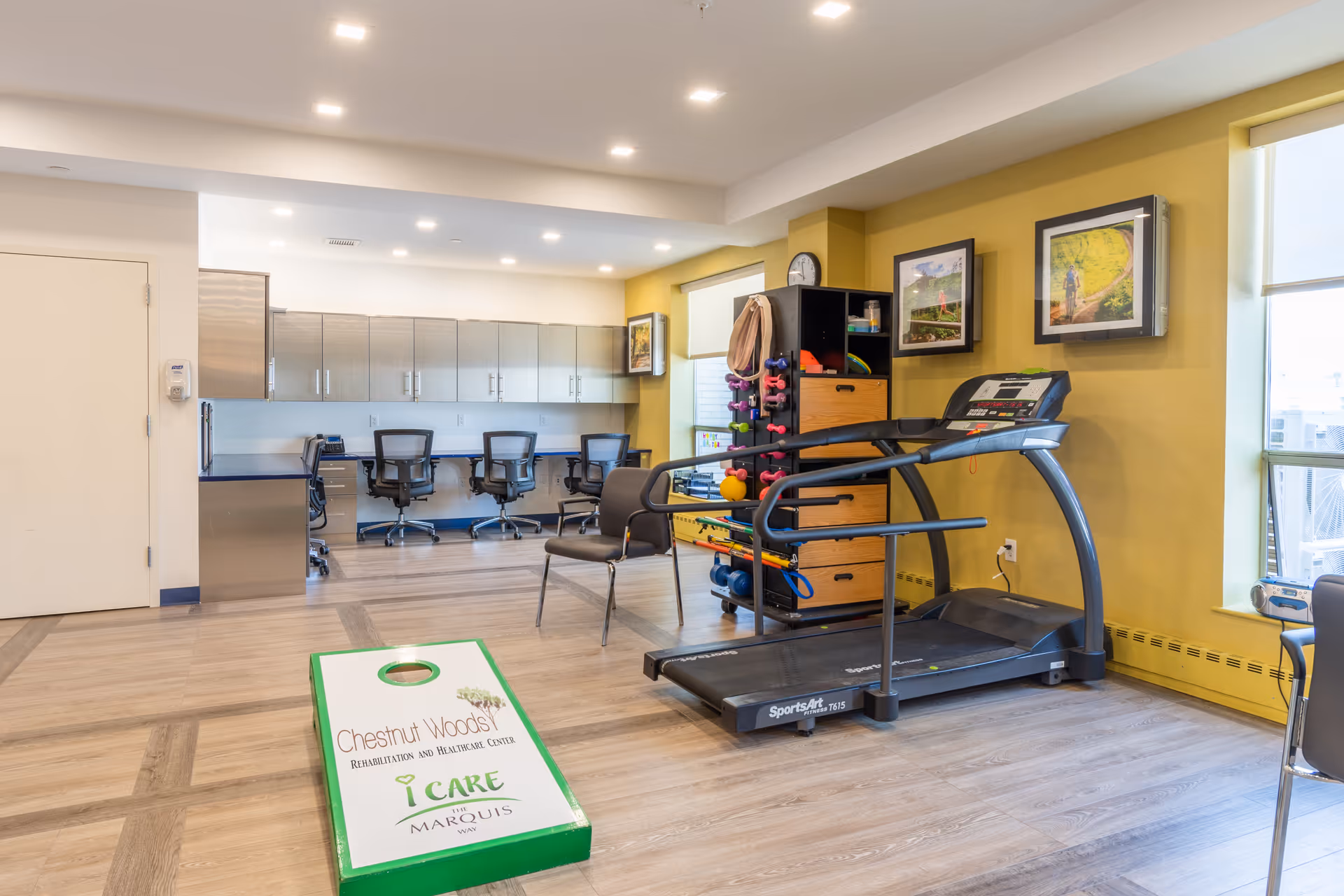 A rehabilitation and healthcare center room featuring a treadmill, exercise equipment rack with weights and therapy tools, a cornhole game board with Chestnut Woods Rehabilitation and Healthcare Center branding, several office chairs and desks with cabinets in the background, and framed pictures on yellow walls.