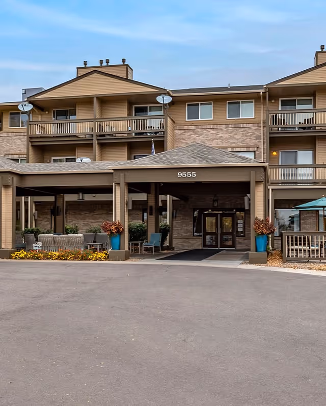 Exterior view of a senior living facility building with a covered entrance labeled 9555. The building has multiple balconies, brick and wood siding, outdoor seating areas with chairs and tables, and potted plants near the entrance.