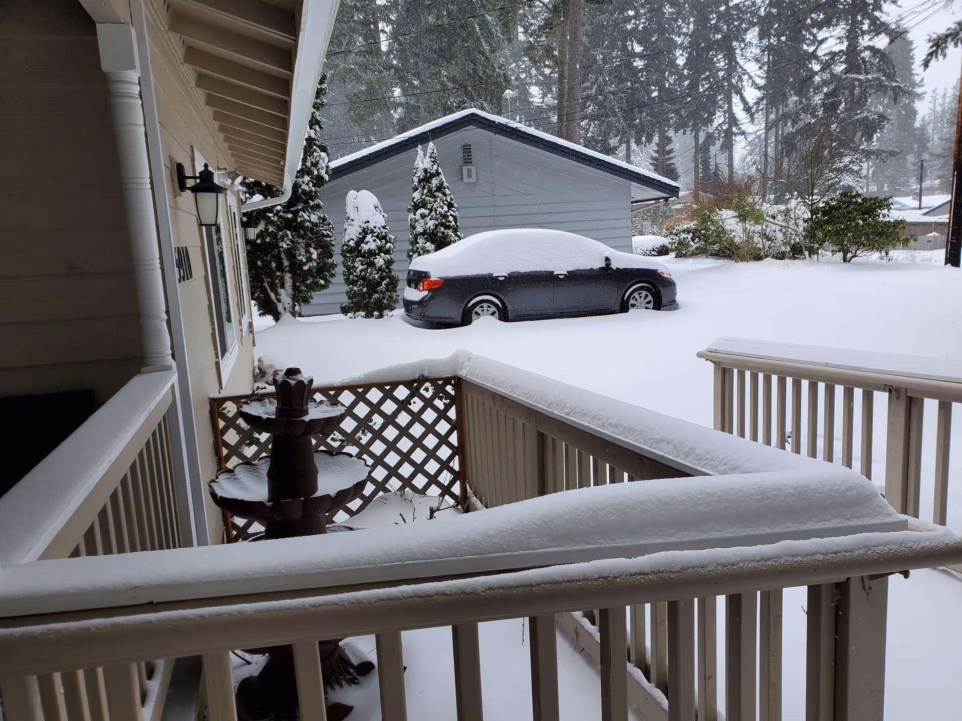 Snow-covered porch railing and patio area with a snow-covered car parked in front of a gray garage. Snow blankets the ground and trees in the background, creating a wintery scene.