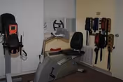 A small rehabilitation room with exercise equipment including a recumbent bike and a standing exercise machine. Several belts or straps are hanging on the wall. The room has beige walls and a window with a closed blind.