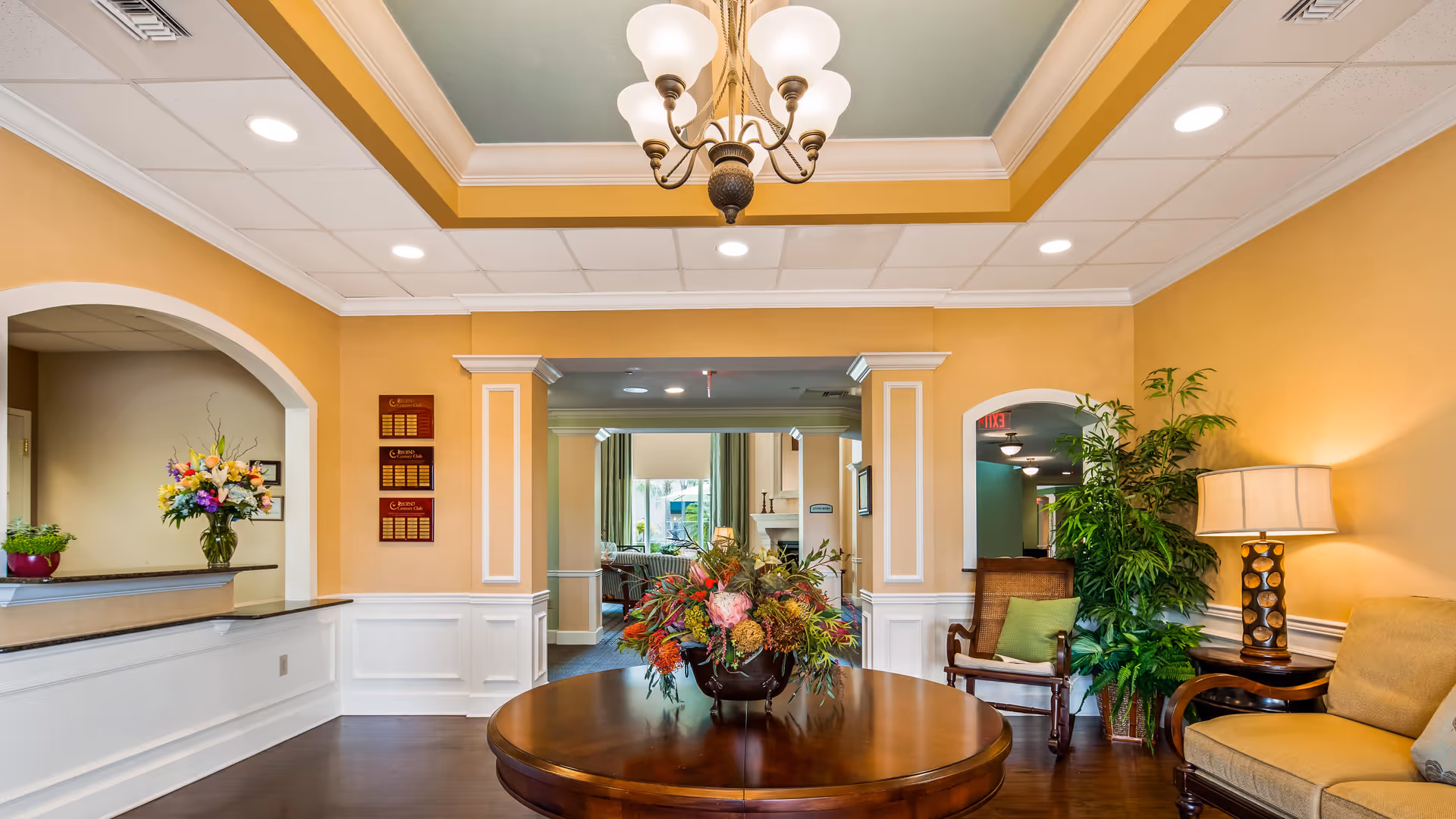 A warmly decorated interior space with yellow walls and white trim, featuring a round wooden table with a large floral arrangement in the center. There is a chandelier hanging from the ceiling, a reception desk with flowers on the left, and seating with a lamp and plants on the right. An open doorway leads to another room with more seating and a fireplace.