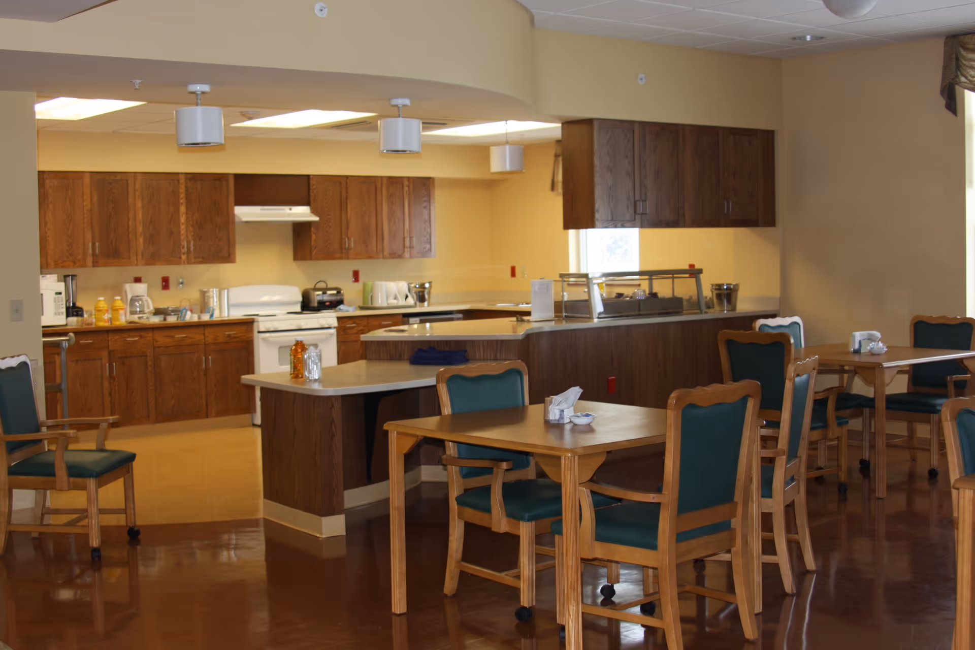 Dining area with wooden tables and green-upholstered chairs facing a serving counter and a kitchen with wooden cabinets.