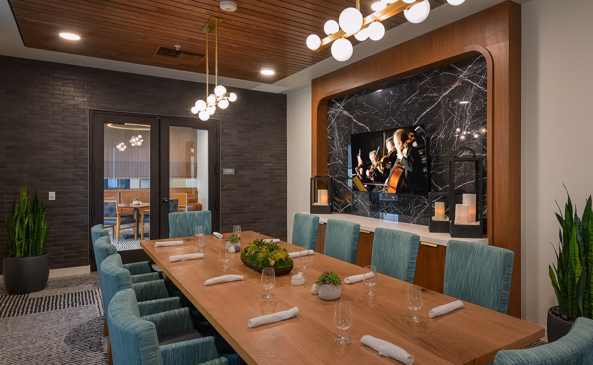 A modern dining room with a long wooden table set with napkins and glasses, surrounded by teal upholstered chairs. The room features a dark brick accent wall with double glass doors, a wooden ceiling with recessed lighting, and a large flat-screen TV mounted on a black marble wall with wooden framing. There are decorative plants and candle lanterns on a white shelf beneath the TV.