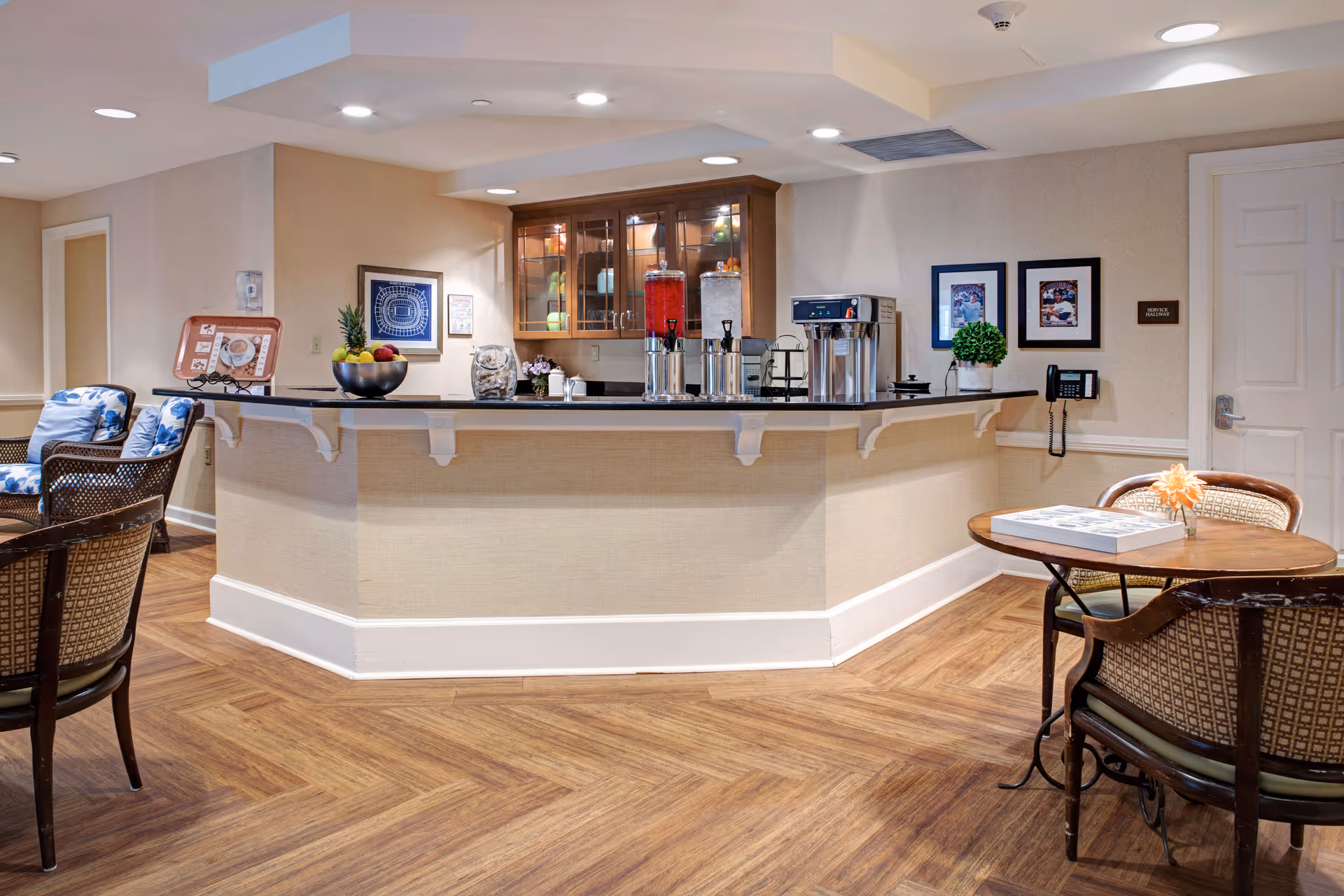 A cozy common area in a senior living facility featuring a counter with beverage dispensers, a coffee machine, and a bowl of fruit. There are chairs with cushions and a small round table with a flower vase and a game on it. The walls are decorated with framed pictures and a telephone is mounted on the wall.