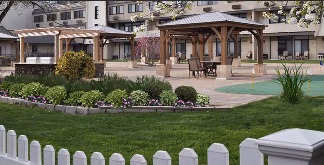 Outdoor courtyard with wooden pergolas, patio seating, landscaped flower beds, a small putting green and a multi-story building in the background.