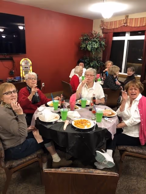 A group of older adults seated around a table eating and drinking in a communal dining room with red walls.