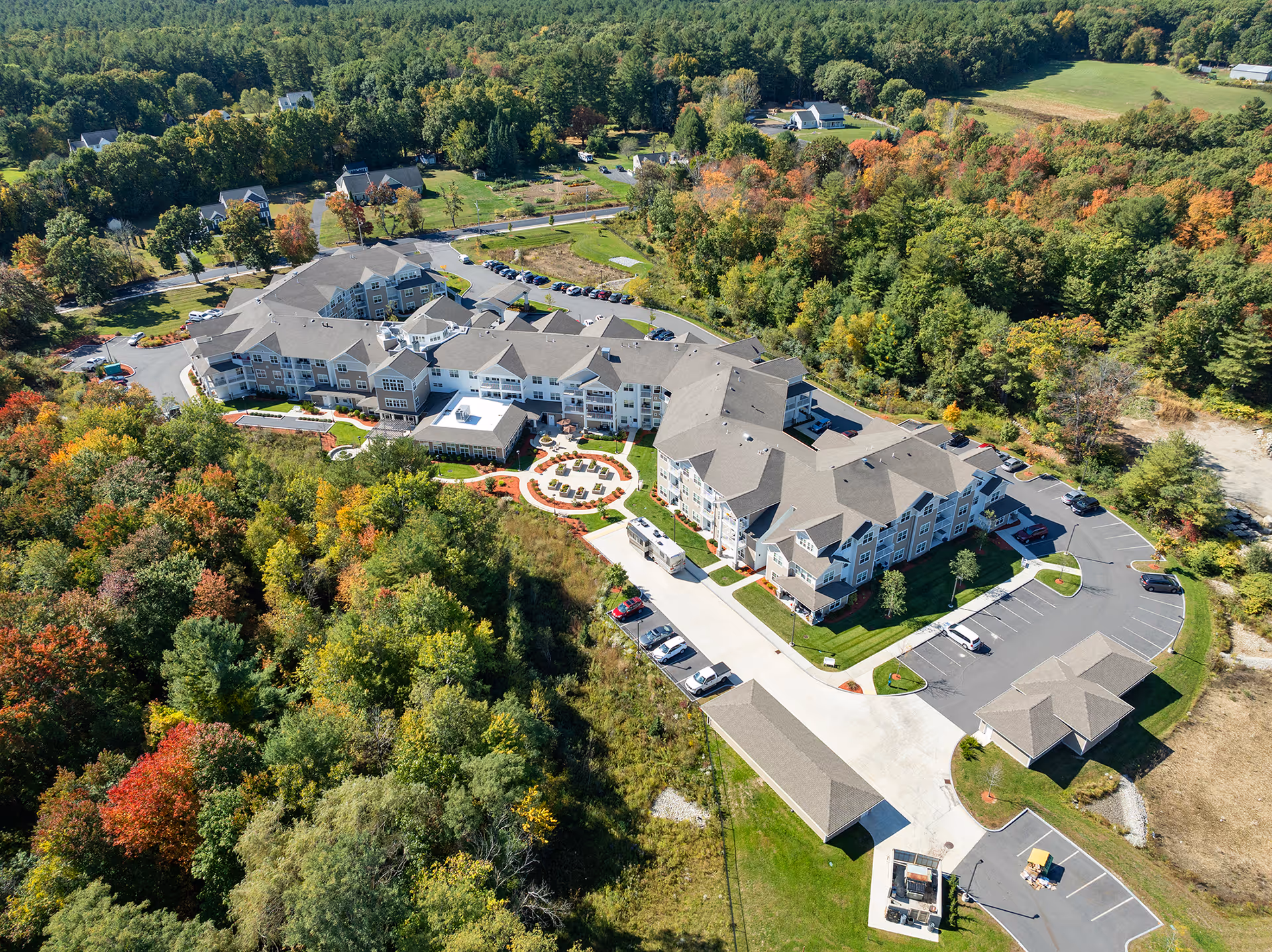 Aerial view of Kennedy Meadows Gracious Retirement Living facility surrounded by dense green and autumn-colored trees, showing a large multi-wing building with parking lots and outdoor seating areas.