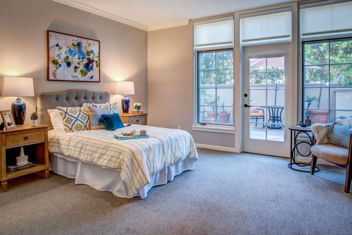 Sunlit bedroom with a tufted bed, patterned pillows, two nightstands with lamps, a chair, and glass doors leading to a patio.
