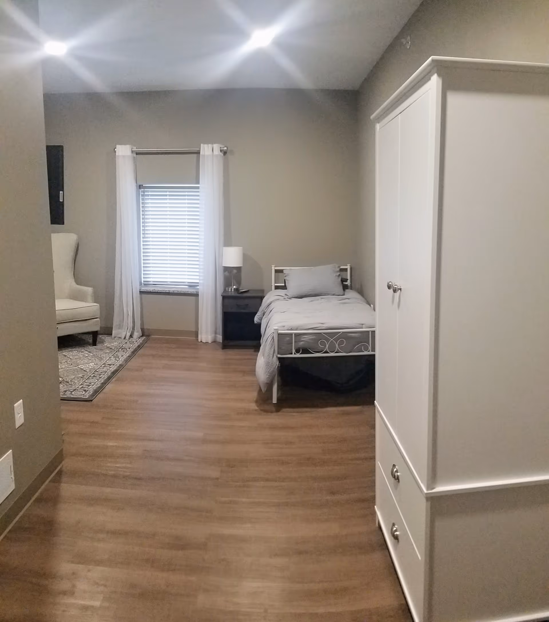 A simple, clean bedroom with a single bed made with gray bedding, a black nightstand with a lamp, a white wardrobe with drawers, a window with white curtains and blinds, and a beige armchair on a patterned rug in the corner.