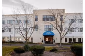 Three-story white senior living facility with blue entrance awning and leafless trees.