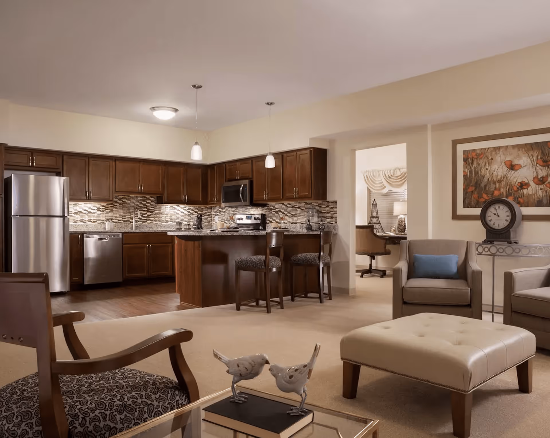 A spacious and well-lit living area with beige walls and carpet. The room features a kitchen with dark wooden cabinets, stainless steel appliances including a refrigerator, dishwasher, and microwave, and a kitchen island with two bar stools. In the living area, there are two beige armchairs with a blue cushion on one, a beige ottoman, and a glass coffee table with two decorative bird figurines and a book. A framed artwork and a clock on a console table are visible on the wall. Through an open doorway, a desk with a chair and a lamp can be seen in an adjacent room.