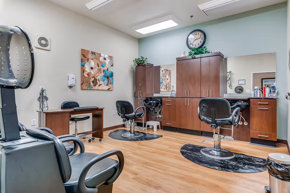 Interior of a senior living facility's hair salon with wooden flooring, two black salon chairs in front of a large mirror and wooden cabinets, a hair dryer chair, a small desk with a chair, a wall clock, and floral artwork on the wall.
