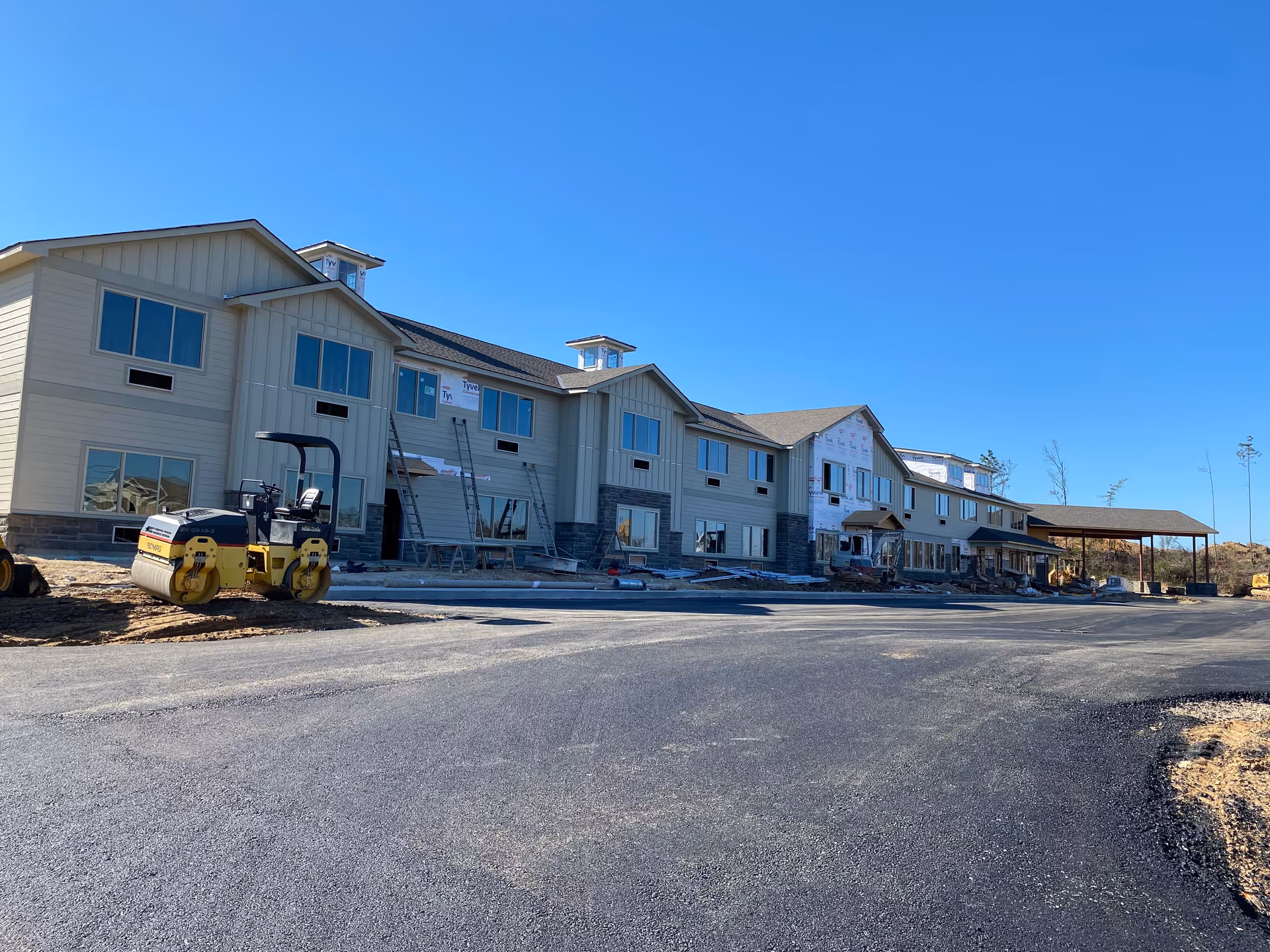 Two-story senior living building under construction with construction equipment and a newly paved driveway under a clear blue sky.