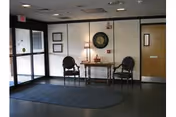 Small lobby/entry area with two chairs flanking a table and lamp beneath a round wall decoration, near glass entrance doors and a wooden interior door.