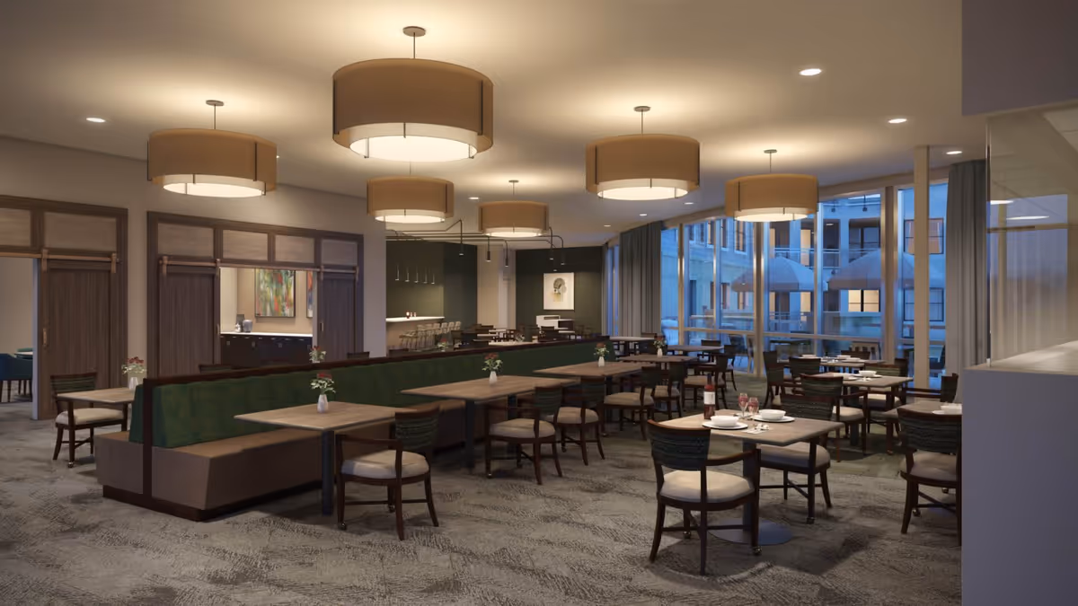 A spacious, modern dining room with multiple tables and chairs arranged neatly. The room features large windows allowing natural light to enter, round ceiling lights with beige lampshades, and a green cushioned bench running along the center. The tables are set with small flower vases, and some have plates, glasses, and a bottle of wine. The decor includes dark wood accents and a painting on the far wall.