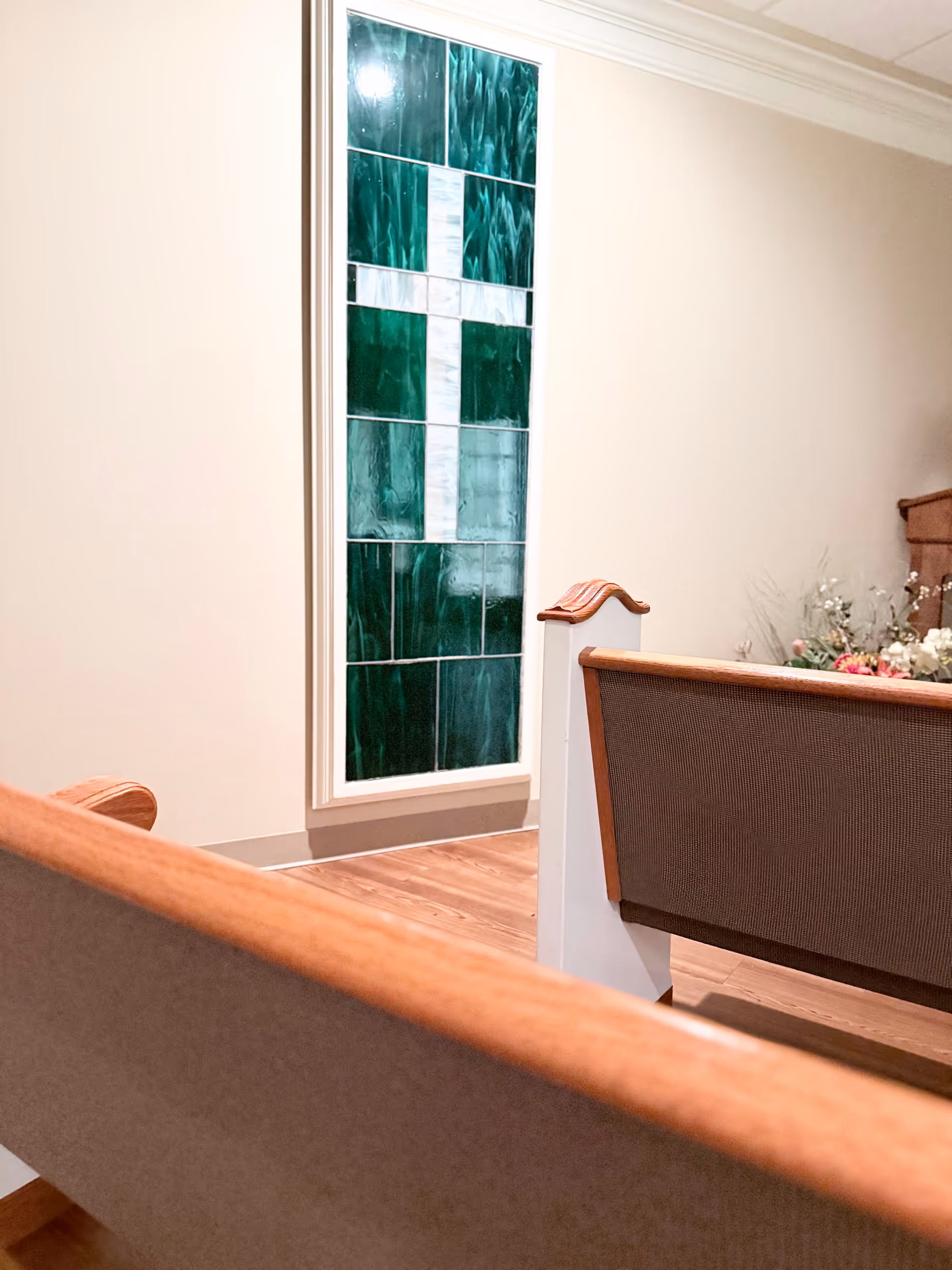 Interior view of a room with wooden pews and a stained glass window featuring a white cross on a green background. There are flowers arranged in the corner, and the walls are painted a light beige color.