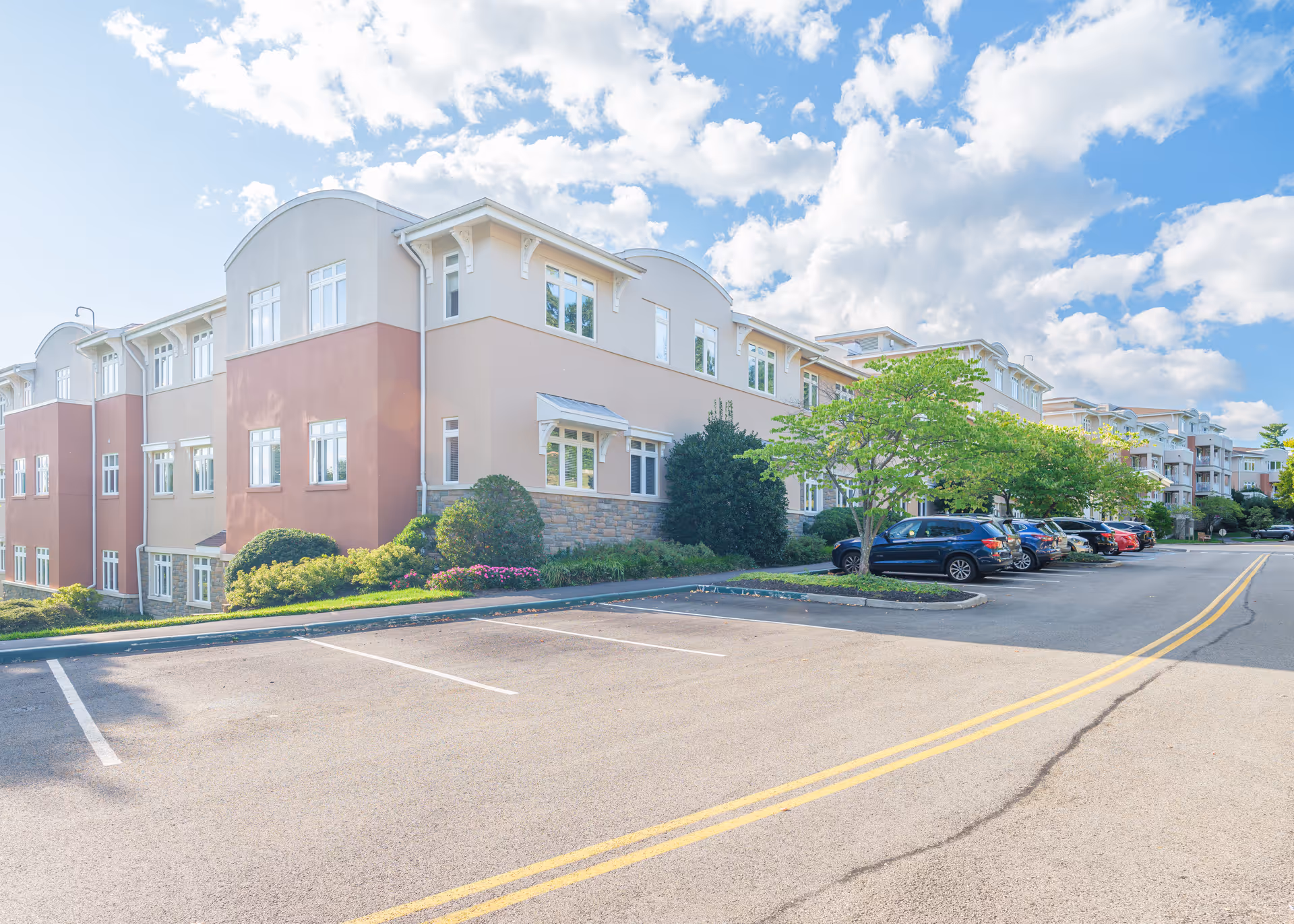 Exterior view of a multi-story residential building with beige and light brown walls, white-framed windows, and a stone base. There is a parking lot in front with several parked cars and a few trees and shrubs along the building. The sky is partly cloudy with blue patches.