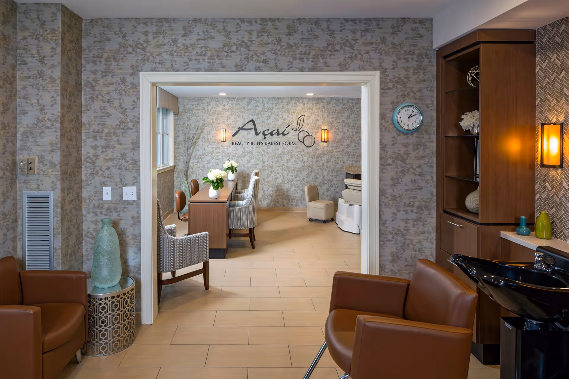 Interior view of a stylish senior living facility room with brown leather chairs, a black wash basin, wooden cabinets, and a decorative side table with a large green vase. Through a doorway, a room with a long table, several chairs, and a wall sign that reads 'Açaí BEAUTY IN ITS RAREST FORM' is visible. The walls have a textured wallpaper and the floor is tiled.