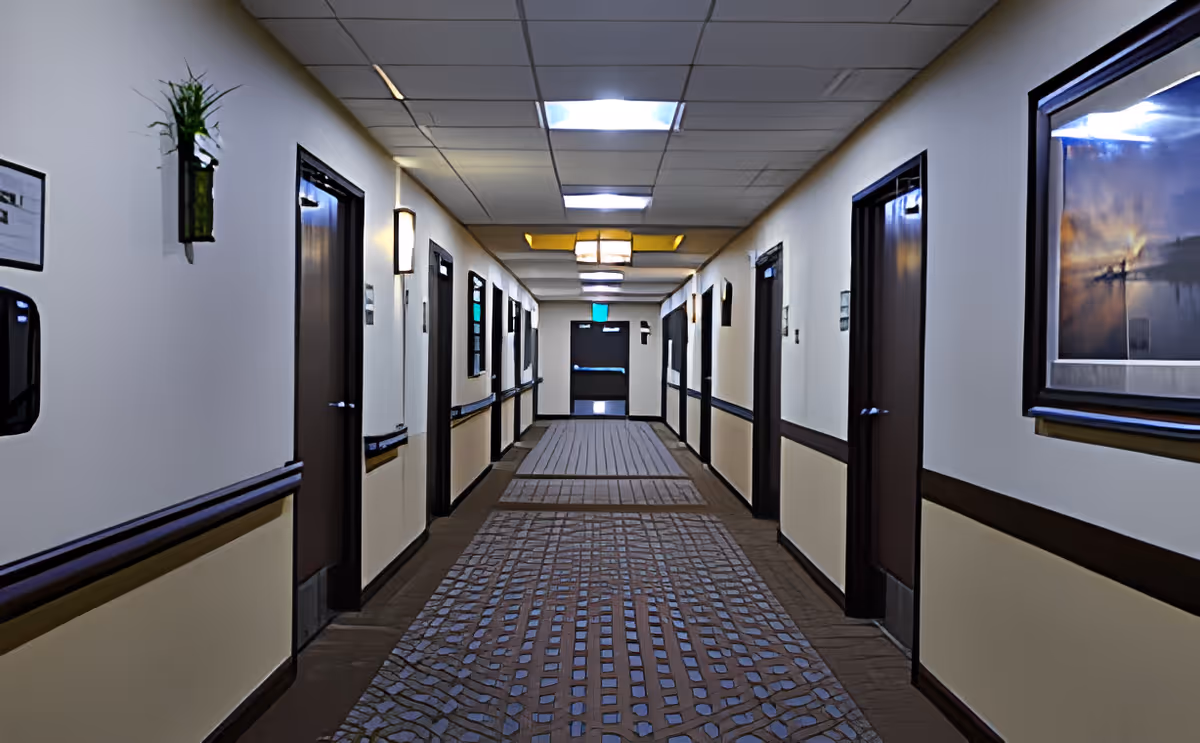 A long, well-lit hallway in an assisted living facility with multiple closed doors on both sides, beige walls with dark brown trim, a patterned carpet, wall-mounted lights, a framed picture on the right wall, and a green exit sign above a door at the far end.