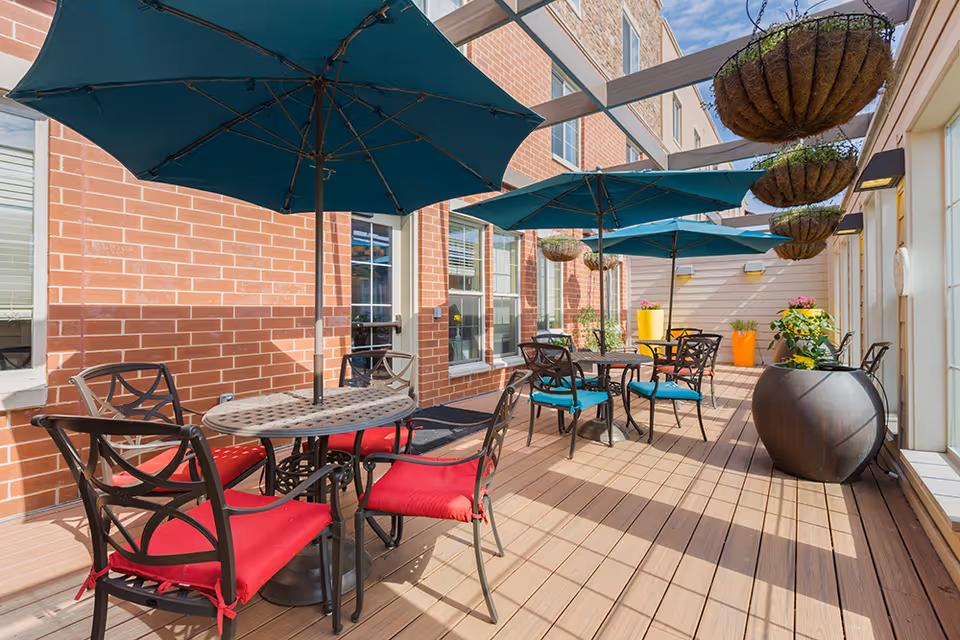 Outdoor patio area with several round metal tables and chairs with red and blue cushions. Large blue umbrellas provide shade over the tables. The patio has a wooden deck floor and is adjacent to a brick building wall with windows. Hanging planters and large potted plants with flowers decorate the space.